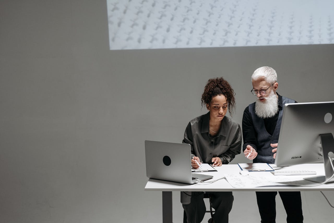 Two colleagues collaborate on a project using laptops and large monitors in a modern office setting.